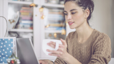 A woman drinking from a mug and using a laptop.