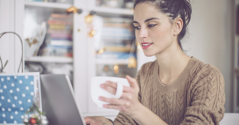 A woman drinking from a mug and using a laptop.